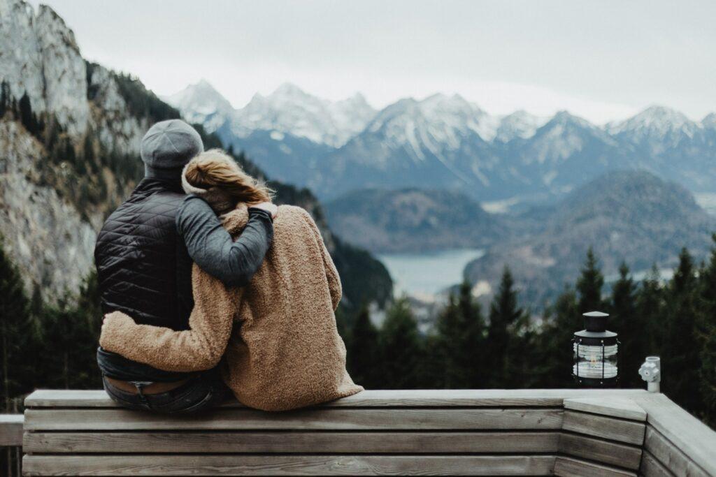 Ein Paar sitzt auf einer Holzbank, umarmt sich und blickt auf schneebedeckte Berge und dichte Wälder des Allgäus. Neben ihnen steht eine Laterne, die eine ruhige, friedliche Stimmung hervorruft.