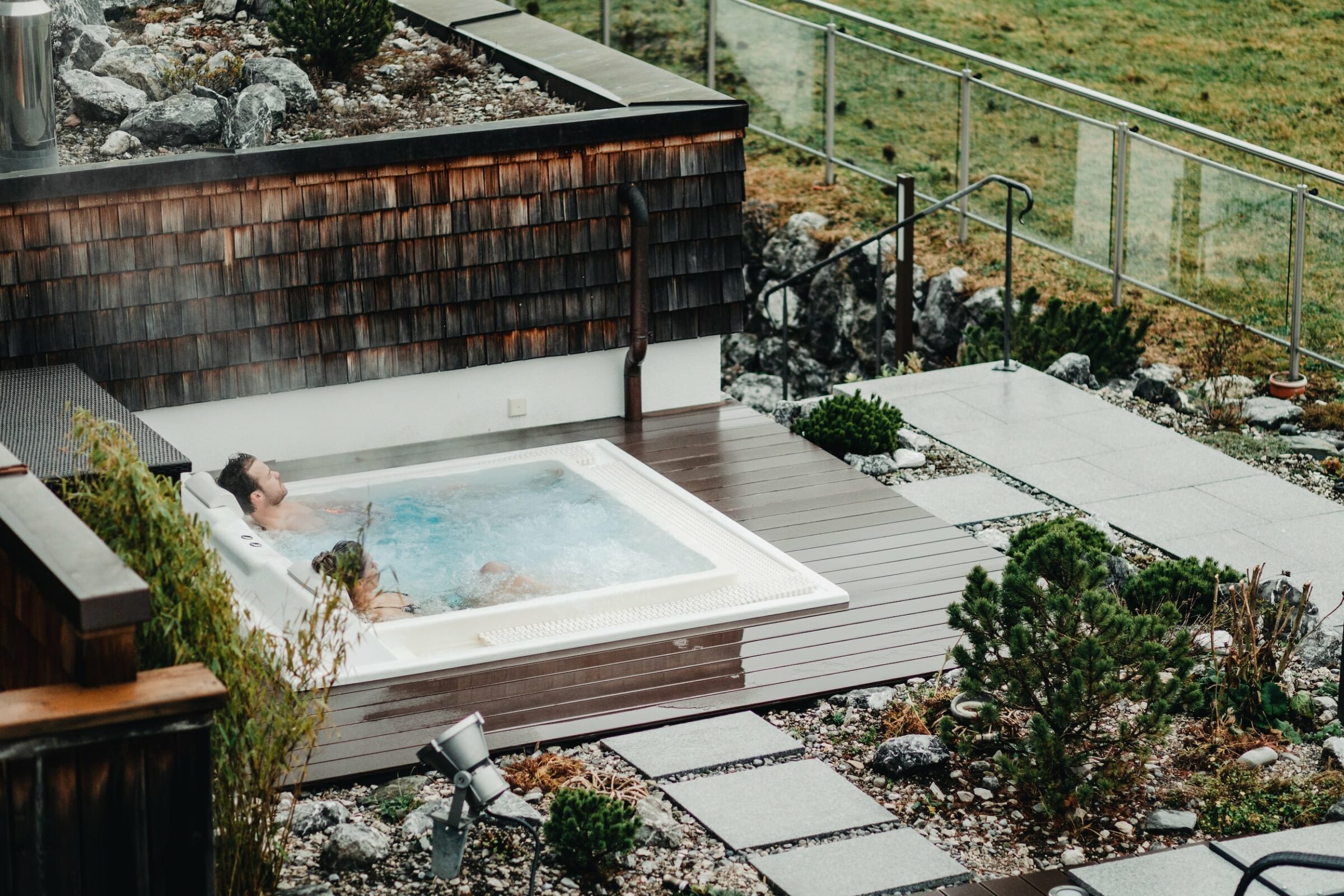 Zwei Personen entspannen sich in einem dampfenden Whirlpool im Freien auf einer Holzterrasse, umgeben von Grün und Steinen, bei bewölktem Himmel und einer friedlichen Atmosphäre im Hotel Das Rübezahl.