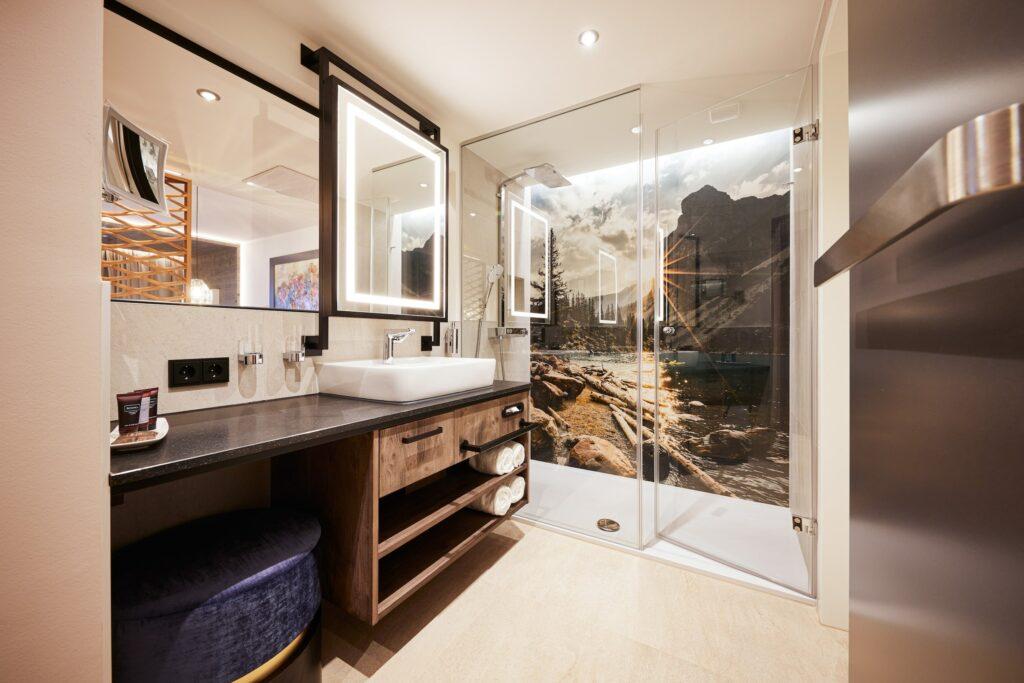 Modernes Badezimmer im nachhaltigen Boutique Hotel Das Rübezahl mit großem Spiegel und beleuchtetem Waschtisch. Eine Glasdusche mit malerischer Berglandschaft sorgt für eine ruhige Atmosphäre.