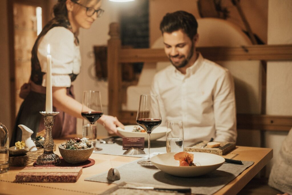 Ein gemütliches Restaurant mit einem Tisch bei Kerzenschein, Weingläsern und Gourmetgerichten. Ein lächelnder Mann sitzt da, während ein Kellner einen Teller serviert. Warme, einladende Atmosphäre.