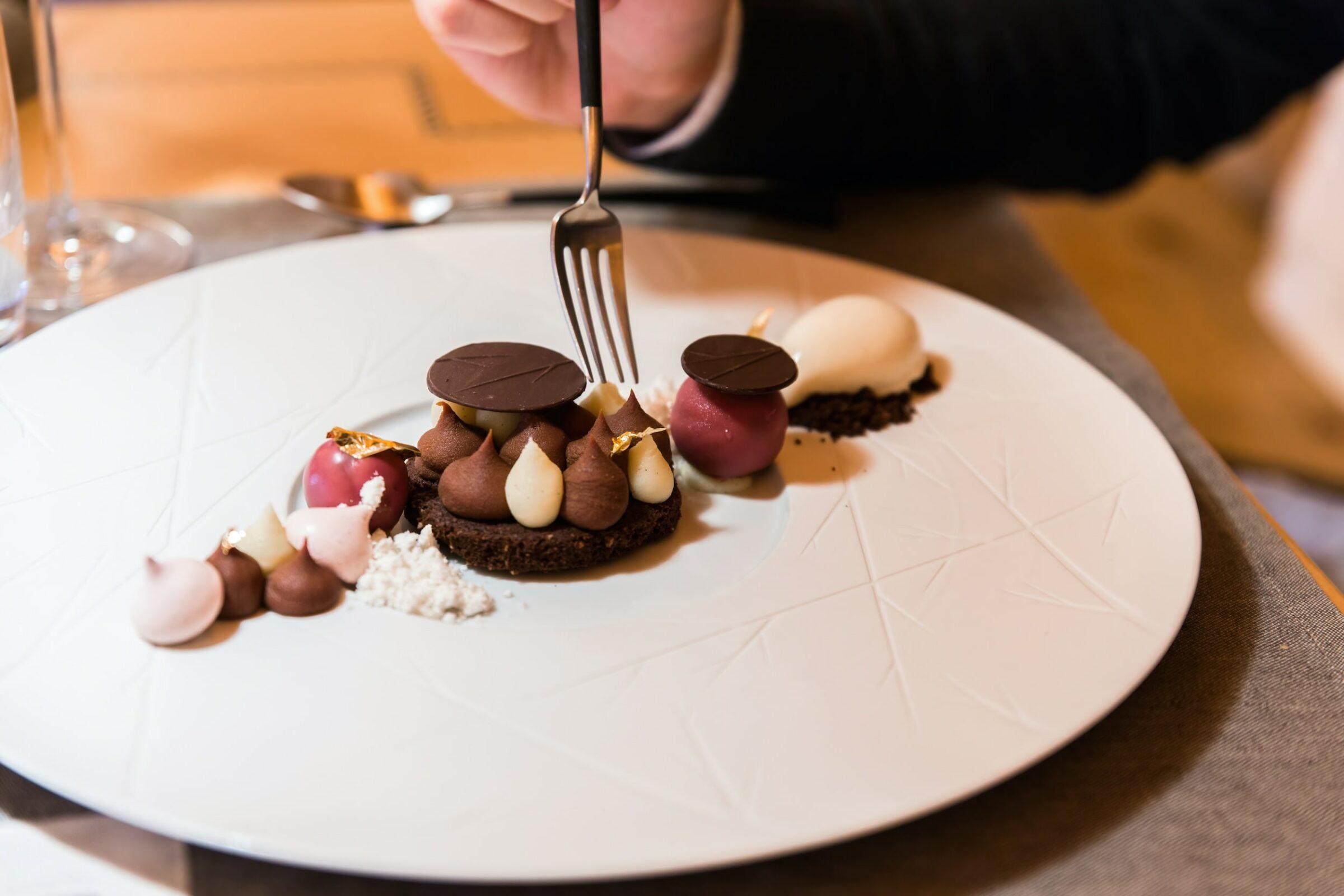 Eine Hand hält eine Gabel über einem Gourmet-Dessert auf einem strukturierten weißen Teller. Das Gericht besteht aus Schokoladenklößchen, roten Kugeln und essbaren Goldakzenten, die eine elegante Präsentation ergeben.