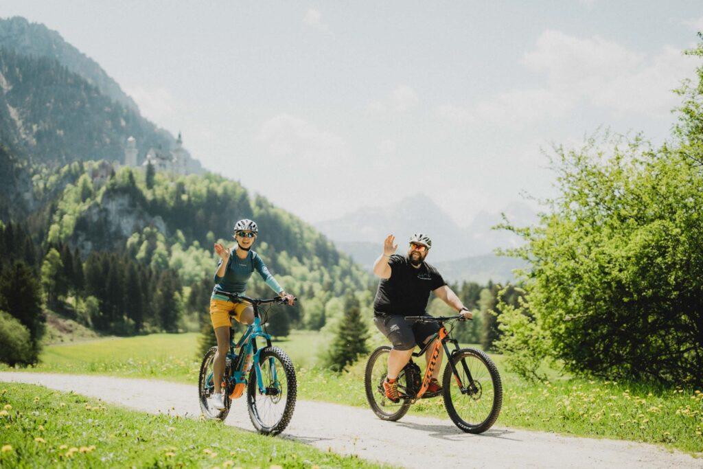 Zwei Radfahrer winken, während sie auf einem malerischen Weg durch üppiges Grün mit Bergen und dem Schloss Neuschwanstein in Bayern im Hintergrund fahren. Die Atmosphäre ist lebhaft und fröhlich.