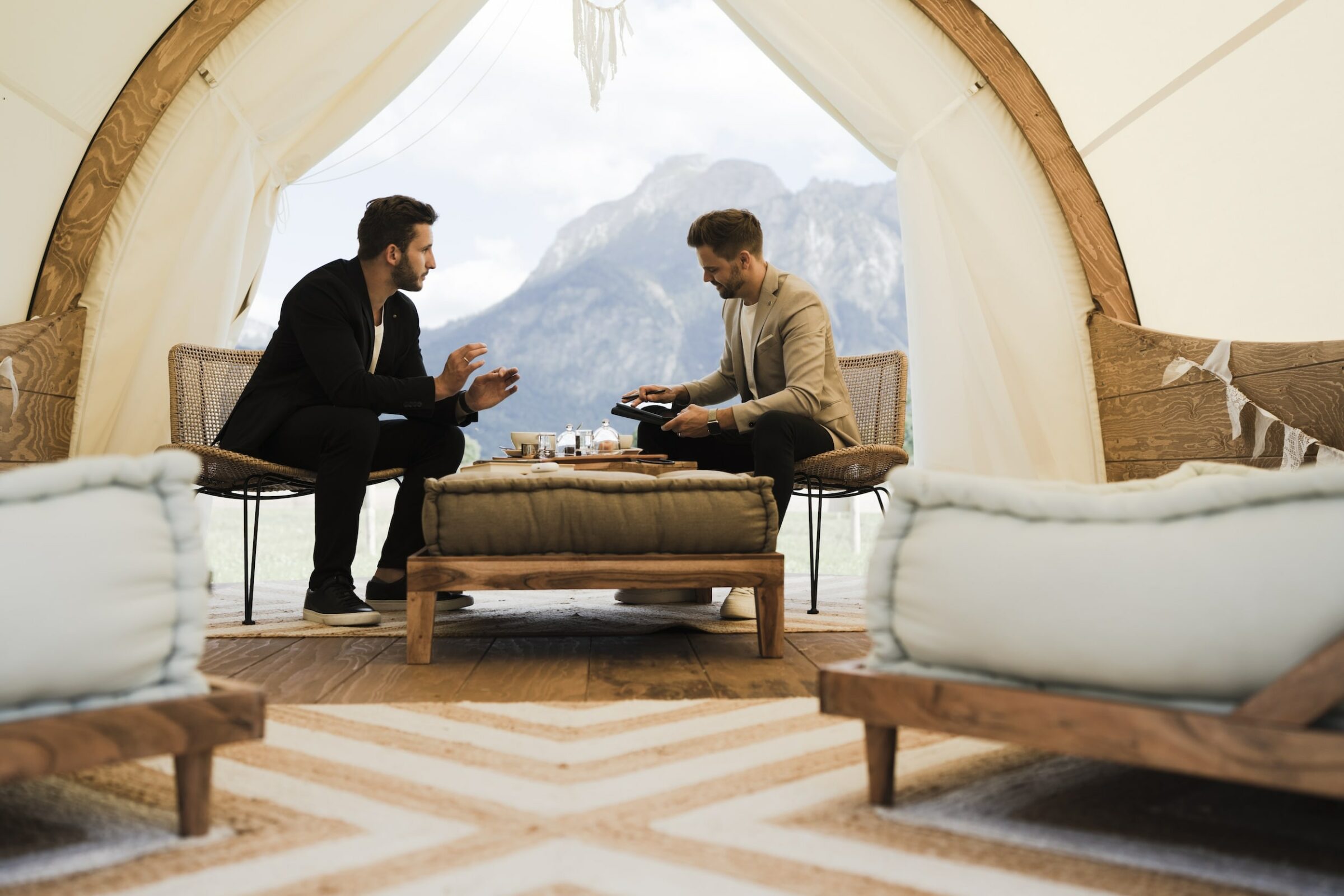 Zwei Männer sitzen in einer gemütlichen, zeltartigen Konstruktion und unterhalten sich. Die rustikale Einrichtung und der Blick auf die Allgäuer Alpen schaffen eine entspannte, einladende Atmosphäre.