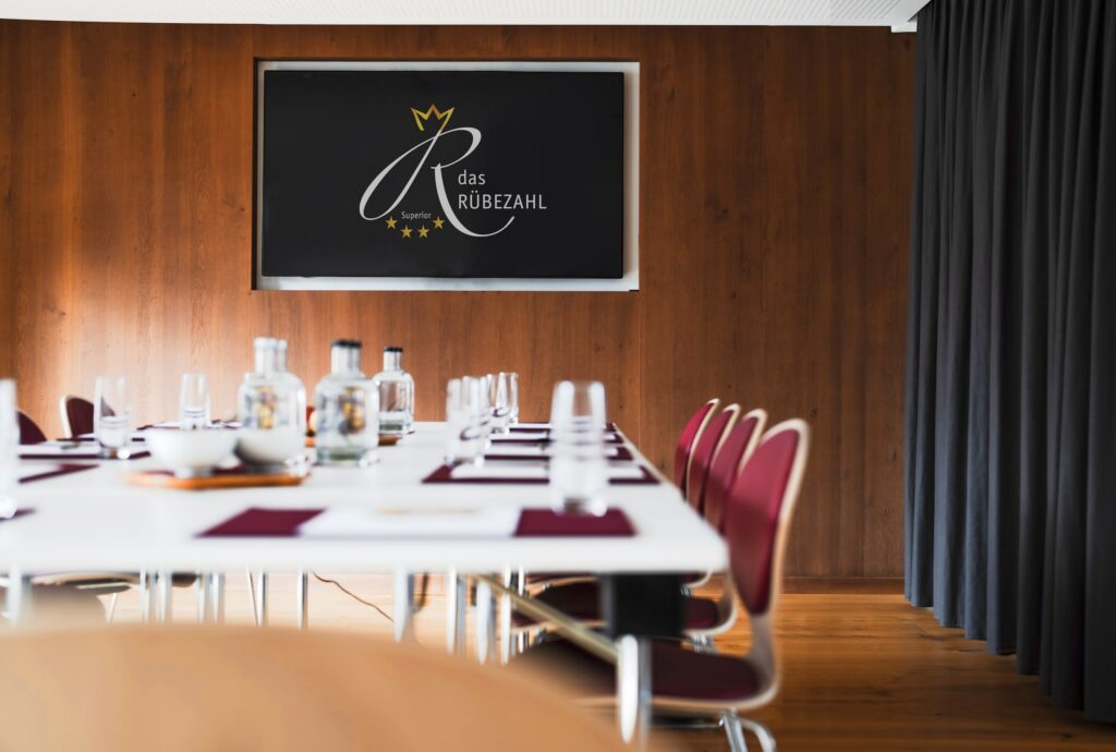 Ein moderner Konferenzraum mit einer Holzwand, auf der ein Bildschirm mit dem „Das Rübezahl“-Logo zu sehen ist. Auf einem Tisch stehen Flaschen, Gläser und rote Stühle.