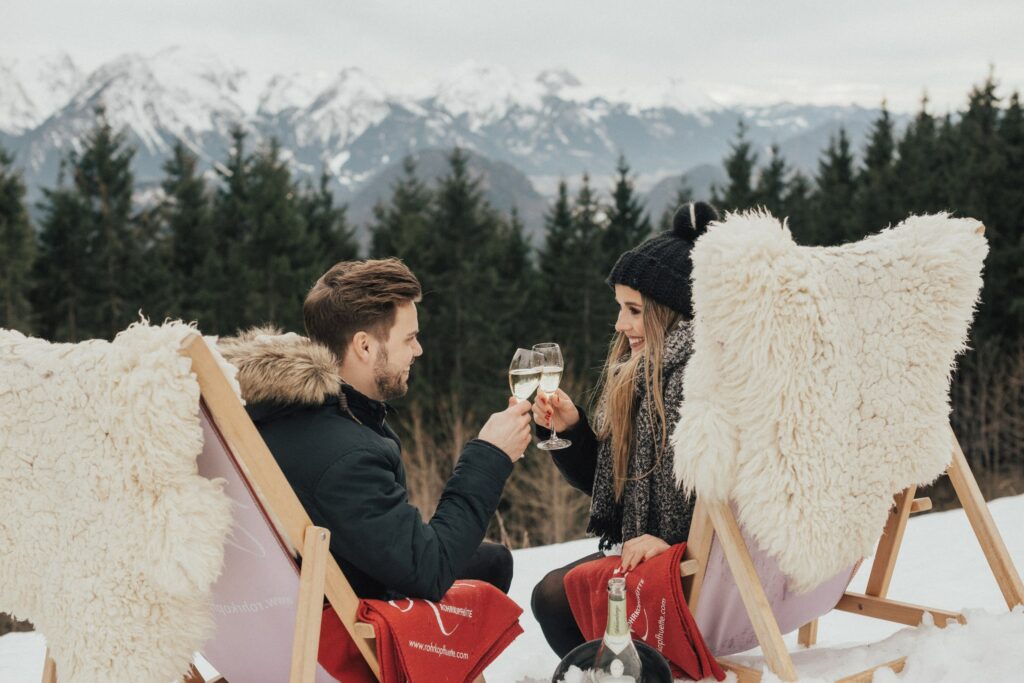 Ein Paar sitzt auf Stühlen mit Felldecken und stößt in einer verschneiten Berglandschaft der Allgäuer Alpen mit Champagner an. Sie wirken gemütlich und fröhlich, hinter ihnen sind Bäume und Berggipfel zu sehen.