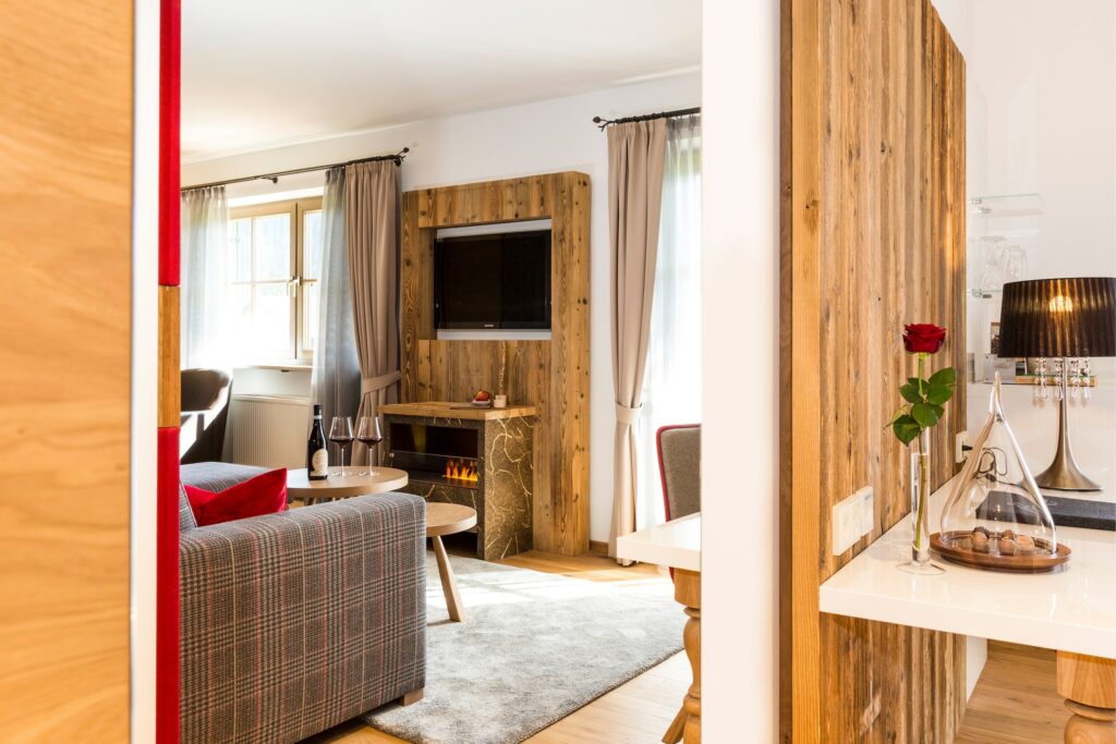 Gemütliches Wohnzimmer in der Schlössl-Suite im bayerischen Hotel Das Rübezahl mit einem karierten Sofa, roten Kissen, einem Holzkamin und einem Fernseher. Eine Blume in einer Vase und Snacks auf einer Theke vermitteln Wärme und Gastfreundschaft.