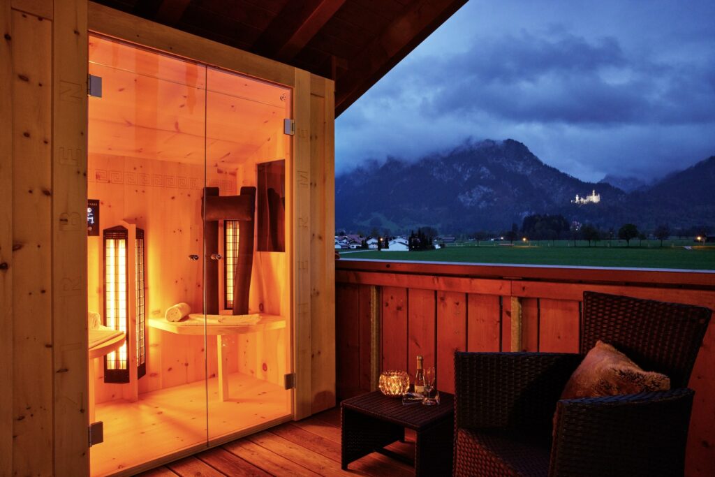 Eine gemütliche Holzsauna strahlt nachts warm auf einem Balkon im Hotel Das Rübezahl im Allgäu, mit Blick auf dunkle Berge und das weit entfernte beleuchtete Schloss Neuschwanstein. Korbstühle tragen zur einladenden Atmosphäre bei.