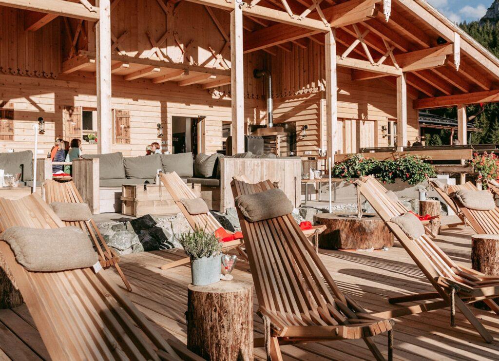 Holzterrasse mit Liegestühlen und gemütlichen Kissen vor der rustikalen Rohrkopfhütte am Tegelberg im Allgäu. Menschen entspannen sich in der warmen, sonnigen Atmosphäre, umgeben von viel Grün.