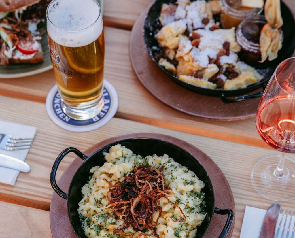 Ein Holztisch mit einem Glas Bier und Roséwein. Auf den Tellern liegen Kässpatzen mit knusprigen Zwiebeln sowie Kaiserschmarrn, der mit Puderzucker bestreut ist. Gemütlich und einladend in der Rohrkopfhütte im Allgäu.