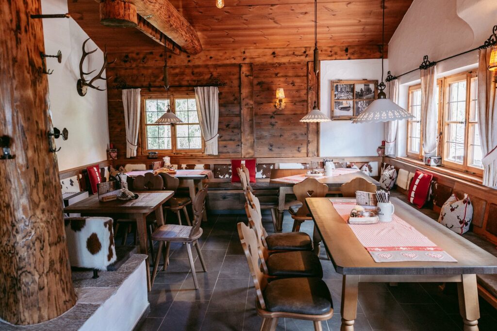 Gemütliches rustikales Esszimmer in der Rohrkopfhütte in den bayerischen Alpen mit Holzwänden und großen Fenstern. Tische mit rot karierten Tischläufern und gepolsterten Stühlen. Warme, einladende Atmosphäre.