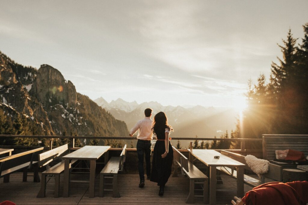 Ein Paar steht auf der Terrasse der Rohrkopfhütte und blickt auf den Sonnenuntergang in den Allgäuer Alpen. Um sie herum stehen Holzbänke, und die Szene strahlt Romantik und Ruhe aus.