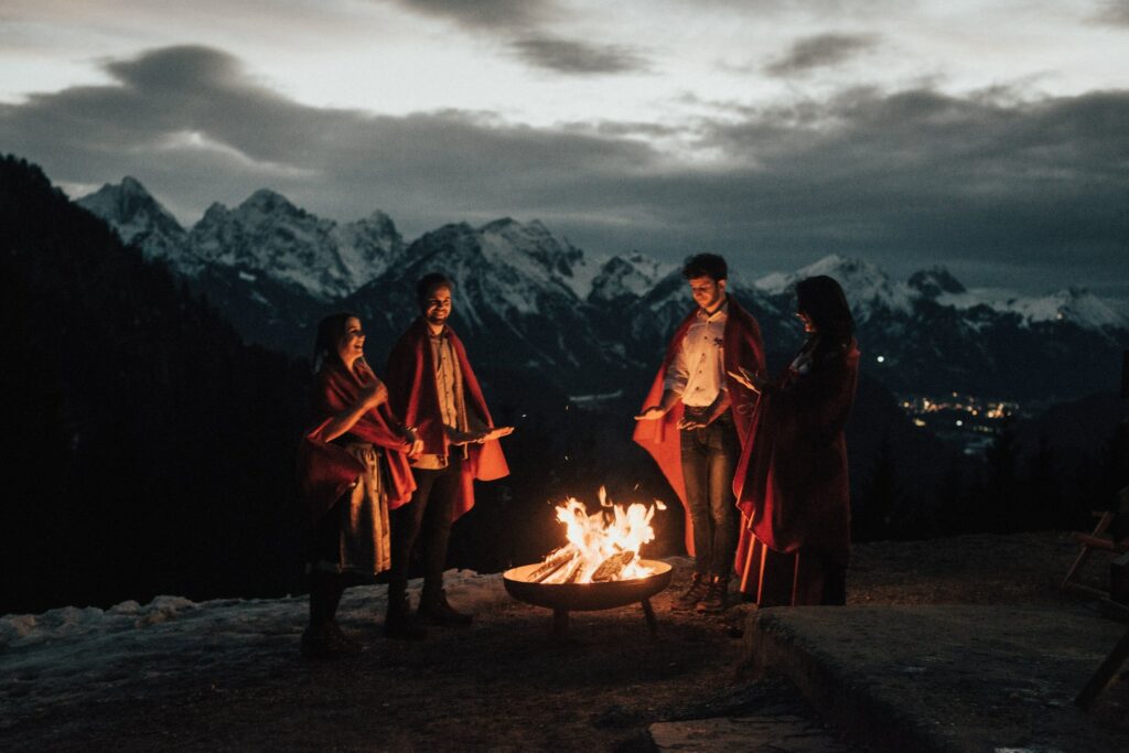 Vier Menschen, in rote Decken gehüllt, sitzen um ein Lagerfeuer herum auf einem Berg mit schneebedeckten Gipfeln und einem dunklen Himmel im Hintergrund.