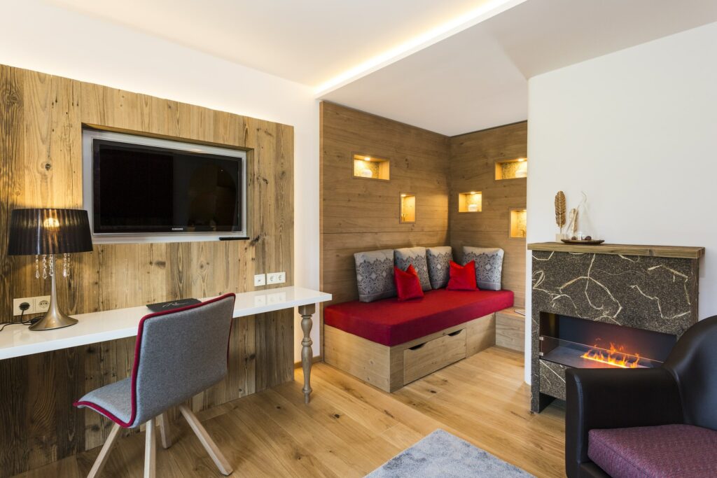 Gemütliches Zimmer im nachhaltigen Hotel Das Rübezahl in Schwangau bei Füssen mit modernem Kamin, rot gepolsterter Holzbank, grauen und roten Kissen, einem an der Wand montierten Fernseher, einem Schreibtisch mit Lampe und stimmungsvoller Beleuchtung. Warme Atmosphäre.