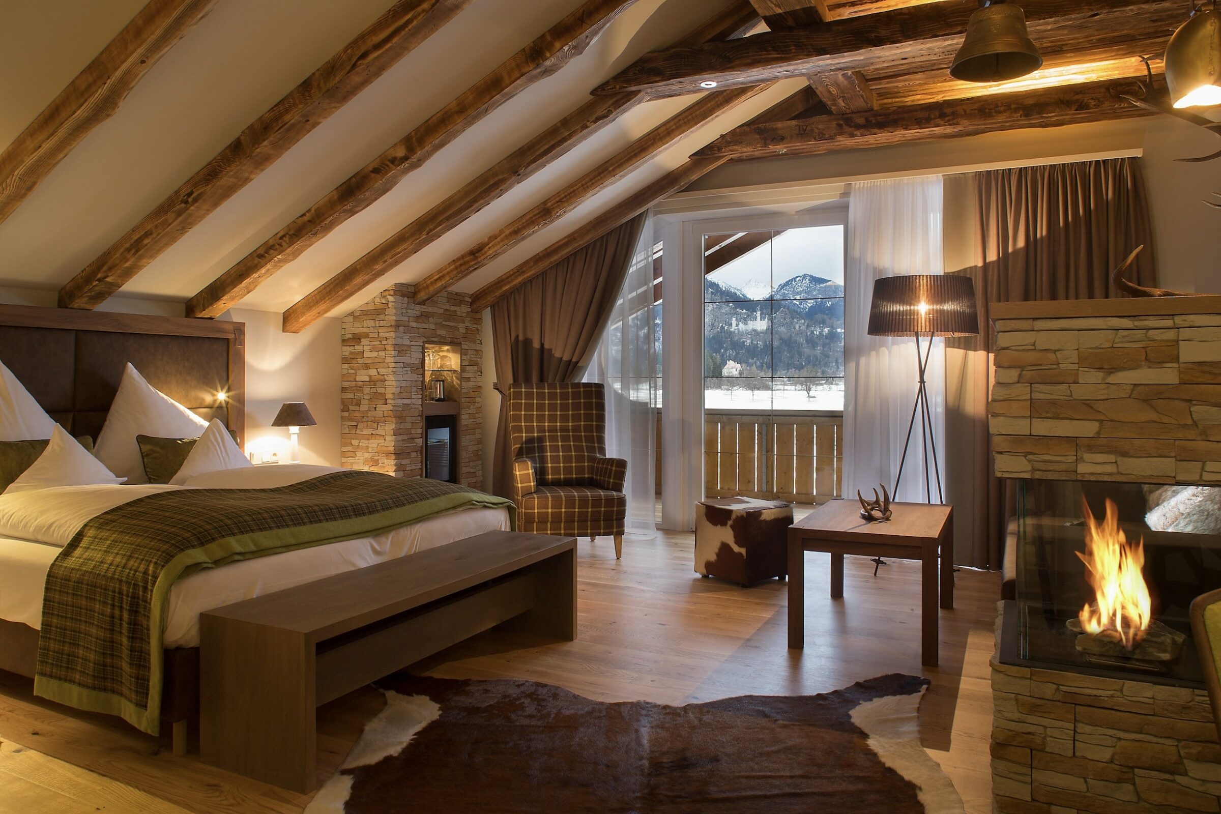 Gemütliche Hotelsuite mit Holzbalken, Steinakzenten, einem großen Bett und einem Kamin. Warme Beleuchtung und ein Fenster geben den Blick auf verschneite Berge frei. Elegante und einladende Atmosphäre.