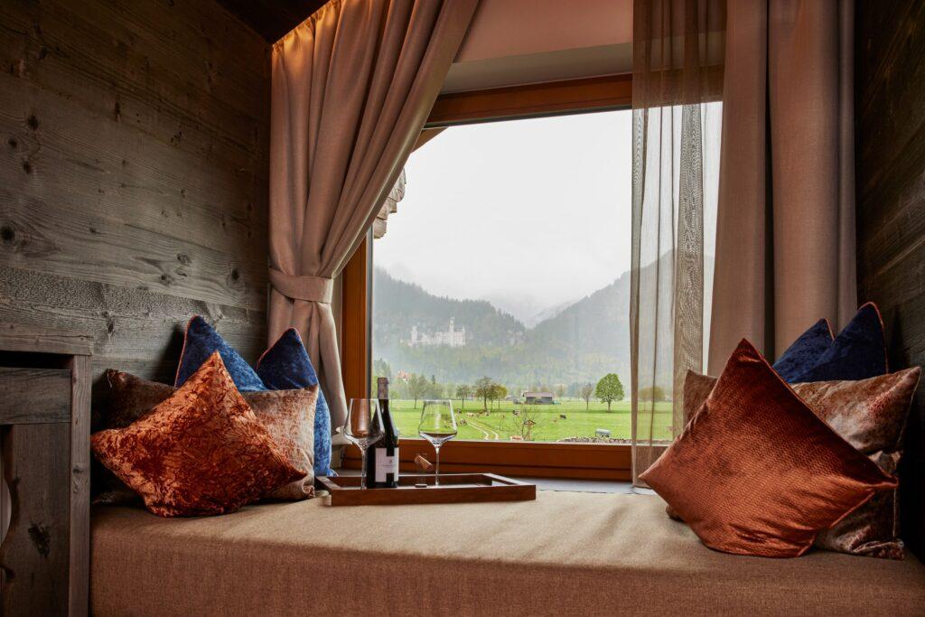 Gemütlicher Fenstersitz mit Samtkissen mit Blick auf neblige grüne Landschaft und entferntes Schloss Neuschwanstein im Allgäu. Weinflasche und Gläser auf Tablett sorgen für einen Hauch von Eleganz.