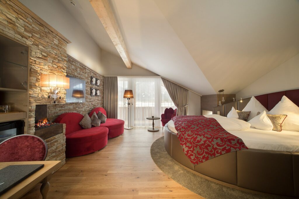 Luxuriöses Hotelzimmer mit schräger Decke und Holzbalken. Mit rundem Bett mit roten Akzenten, Steinkamin, roten Sofas und großen Fenstern mit transparenten Vorhängen, die eine warme, gemütliche Atmosphäre schaffen.