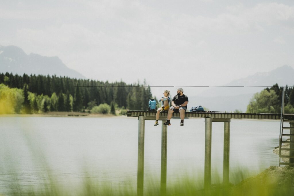 Zwei Menschen sitzen auf einem Holzsteg mit Blick auf einen ruhigen Bergsee in Bayern nahe des nachhaltigen Hotels Das Rübezahl, umgeben von üppigen Wäldern und fernen Bergen, und vermitteln eine friedliche, entspannte Stimmung.