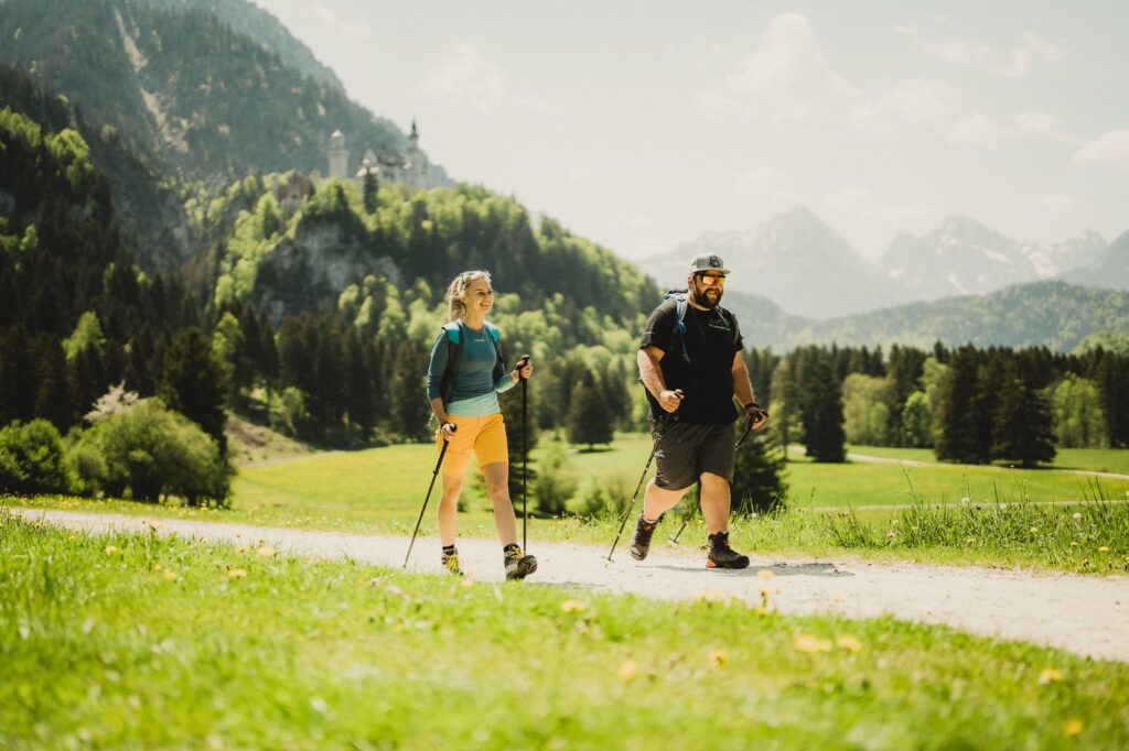Zwei Wanderer mit Wanderstöcken genießen einen sonnigen Tag auf einem üppig grünen Wanderweg nahe des Hotel Das Rübezahl, umgeben von Bergen und Bäumen, und schaffen so eine friedliche und abenteuerliche Szene. Im Hintergrund ist das bayerische Königsschloss Neuschwanstein zu sehen.