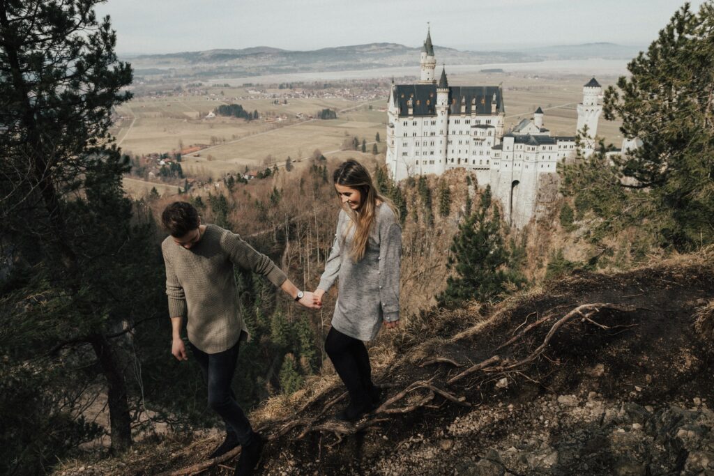 Ein Paar hält Händchen, während es einen bewaldeten Hügel hinaufgeht, im Hintergrund das romantische Schloss Neuschwanstein in Bayern. Die Szene vermittelt Romantik und Abenteuer.
