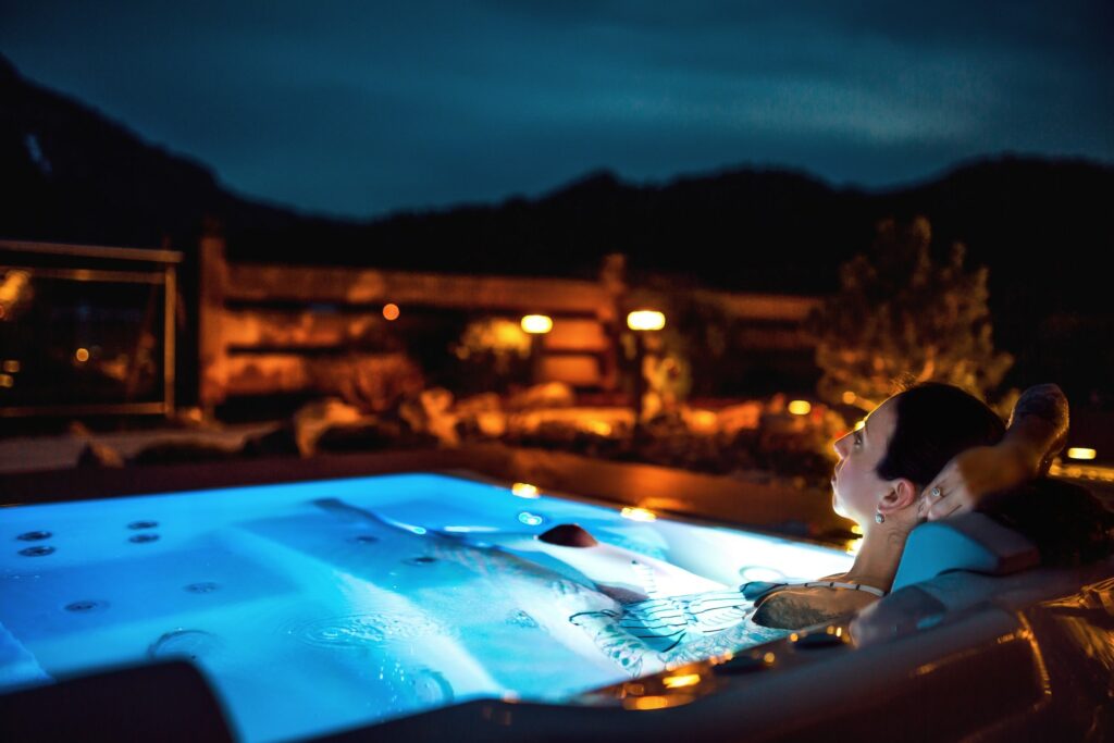 Eine Person entspannt sich nachts in einem leuchtenden Whirlpool im nachhaltigen Boutique Hotel Das Rübezahl im Allgäu, umgeben von einer ruhigen Außenumgebung mit sanften, warmen Lichtern und silhouettenhaften Bergen. Ruhige und friedliche Atmosphäre.