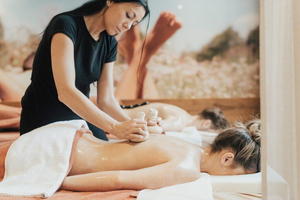 Ein Massagetherapeut in der Beauty Alm des romantischen Hotel Das Rübezahl in Bayern verwendet Kräuterkompressen bei einer entspannten Frau, die mit dem Gesicht nach unten auf einem Massagetisch liegt, und schafft so eine ruhige und beruhigende Spa-Atmosphäre.