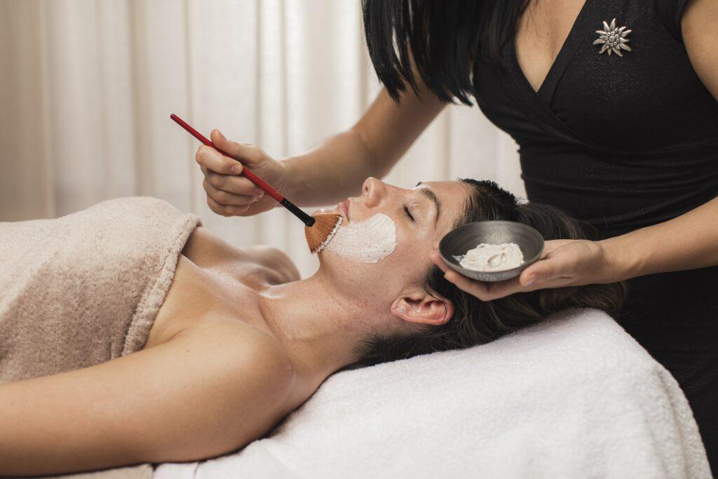 Eine Frau entspannt sich auf einer Wellnessliege in der Beauty Alm des nachhaltigen Boutique Hotels Das Rübezahl in Bayern. Sie ist in ein Handtuch gewickelt, während eine Kosmetikerin ihr mit einem Pinsel eine weiße Gesichtsmaske aufträgt. Die Umgebung ist ruhig und beruhigend.