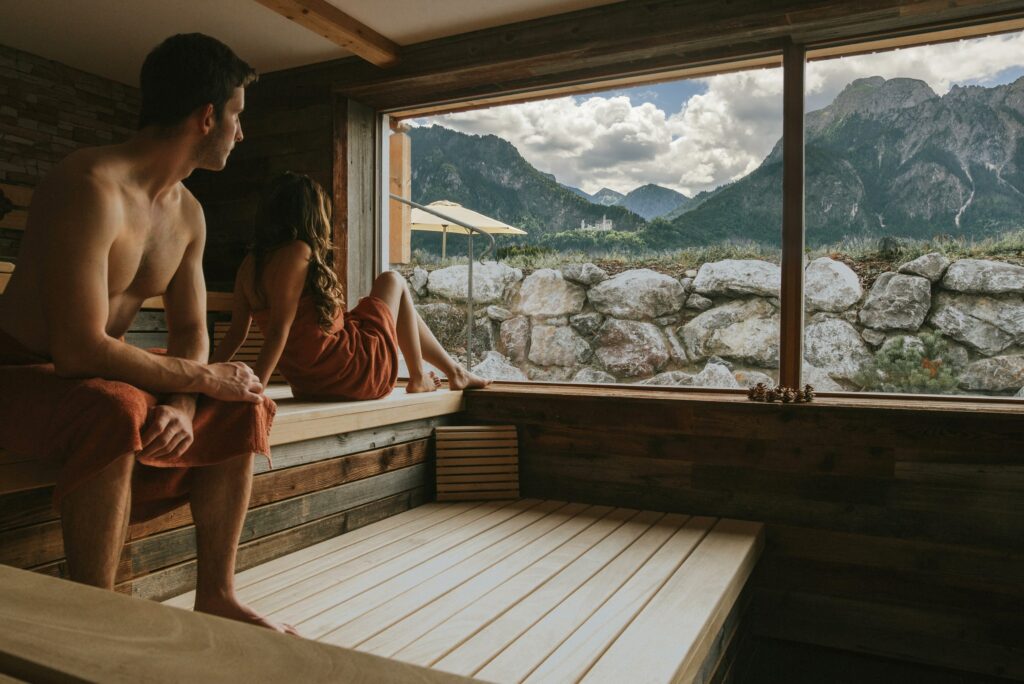 Ein Mann und eine Frau sitzen in Handtüchern in der Holzsauna des Hotel Das Rübezahl in Schwangau und blicken aus einem großen Fenster auf eine malerische Berglandschaft mit dem Schloss Neuschwanstein. Die Atmosphäre ist friedlich und besinnlich.