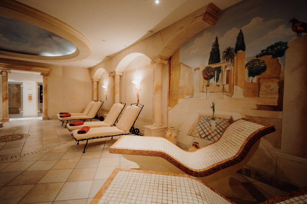 Luxuriöser Wellnessraum der Therme Romana im Hotel Das Rübezahl mit gefliesten Liegen, roten Handtüchern und sanfter Beleuchtung im römischen Stil. Ein Wandgemälde, das eine sonnige Mittelmeerszene darstellt, unterstreicht die entspannende Atmosphäre.