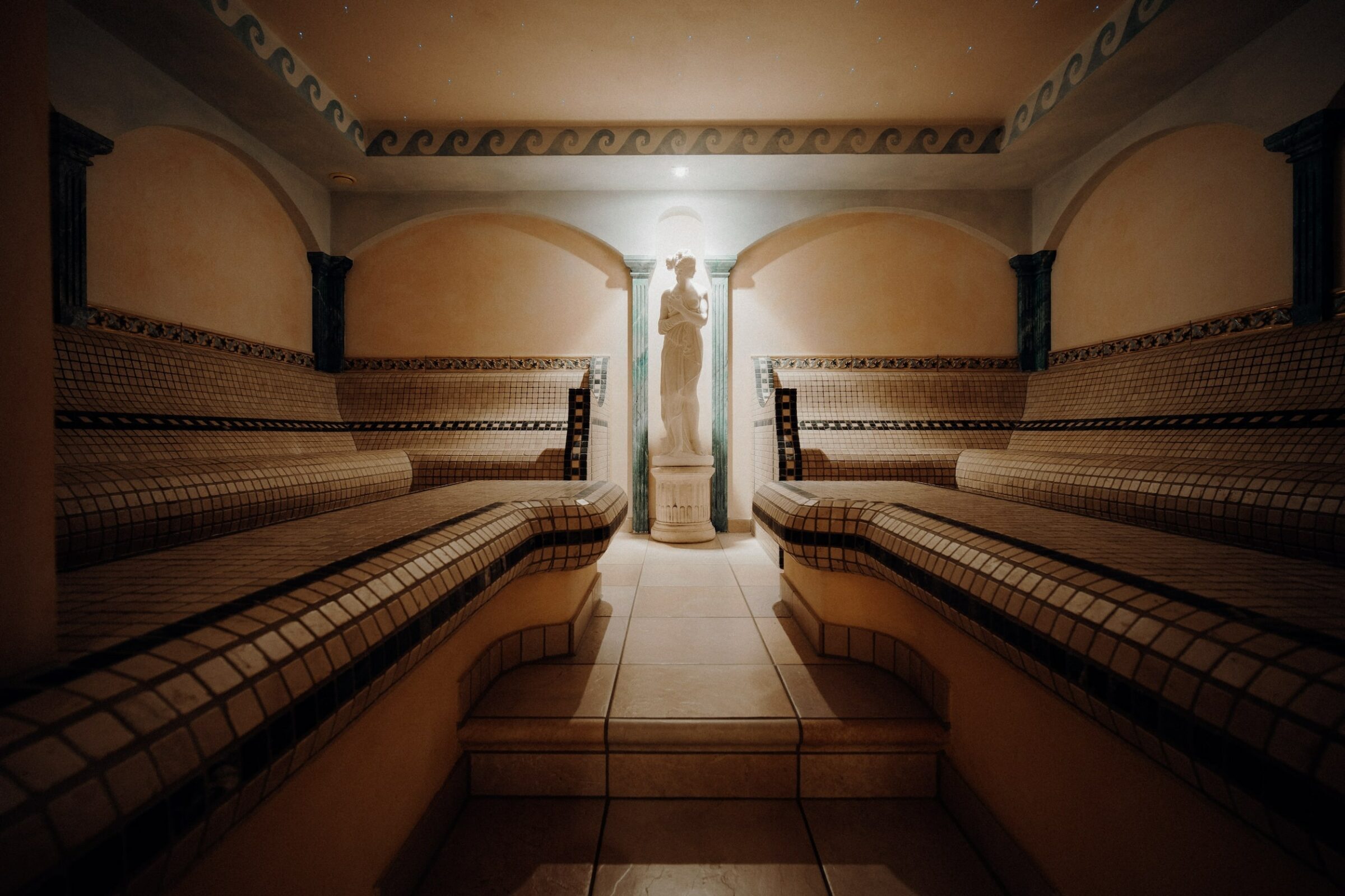 Luxuriöser, gefliester Raum in ther Therme Romana des nachhaltigen Romantikhotels Das Rübezahl mit eleganten Mosaikbänken an den Wänden und einer klassischen Statue in der Mitte am Ende. Sanfte, warme Beleuchtung schafft eine ruhige, spa-ähnliche Atmosphäre.