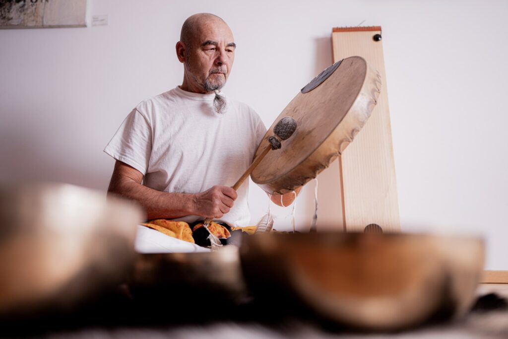 Yoga-Meister Isvan vom Hotel Das Rübezahl in Bayern in einem weißen Hemd spielt ruhig sitzend auf einer großen Handtrommel. Der Raum ist sanft beleuchtet und enthält verschiedene Musikinstrumente, die eine ruhige Atmosphäre schaffen.