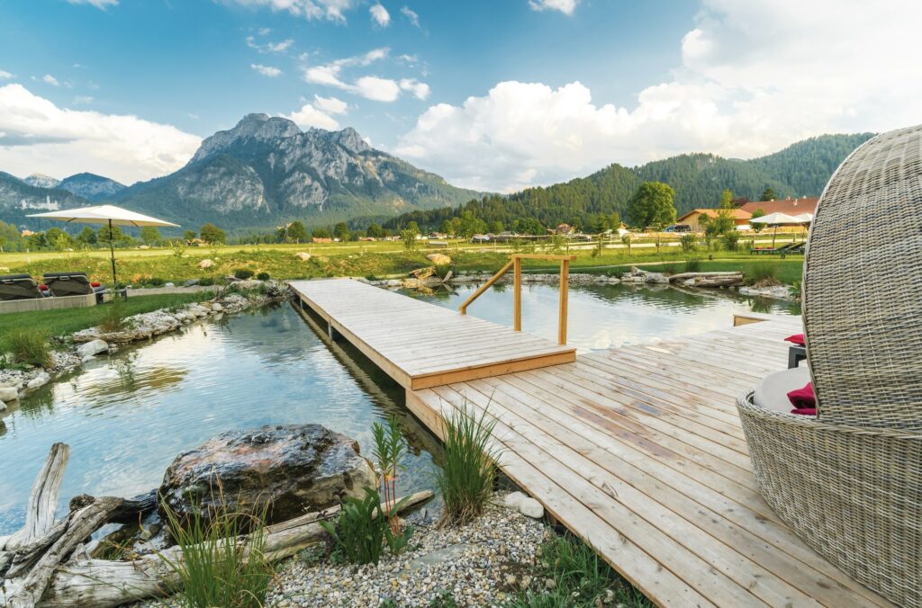 Eine ruhige Landschaft im Allgäu mit einem Holzsteg, der sich über einen klaren Naturschwimmteich erstreckt, umgeben von grasbewachsenen Feldern und Bergen. In der Nähe steht ein Korbstuhl, der Ruhe ausstrahlt. Das ist der Wellnessgarten im Romantic Hideaway Hotel Das Rübezahl