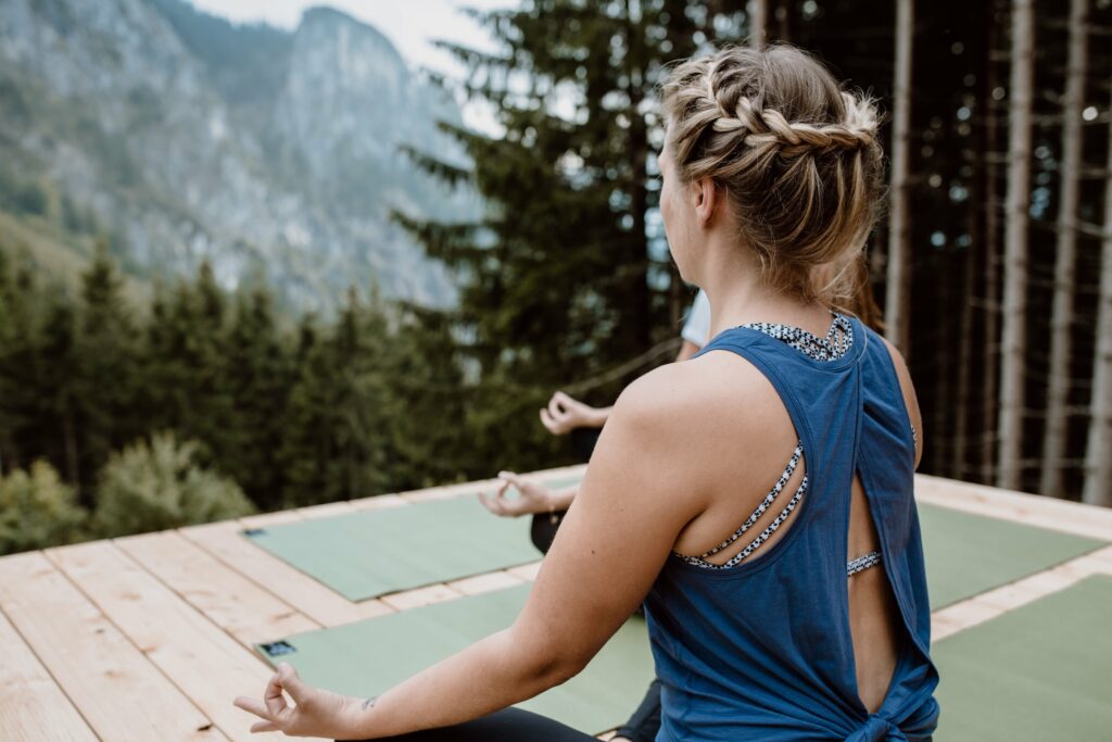 Eine Person mit geflochtenen Haaren, die ein blaues Tanktop trägt, meditiert im Freien auf der Holzterrasse der Rohrkopfhütte im Allgäu mit Yogamatten, umgeben von Bäumen und Bergen. Ruhige Atmosphäre.