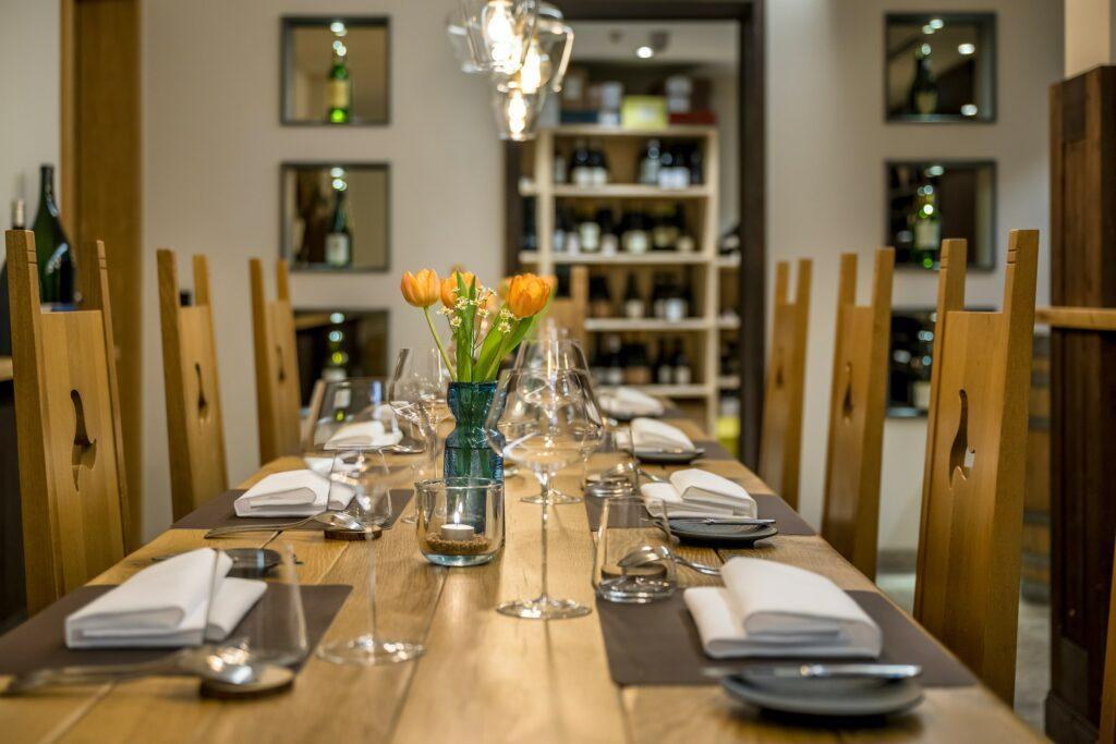 Elegantly laid dining table for dinner at the sustainable wellness hotel Klosterhof in Bavaria with wooden chairs, white napkins, wine glasses and orange tulips in a vase. Shelves with bottles behind them create a cozy, upscale atmosphere.