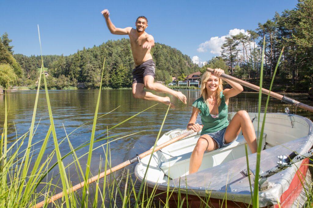 Eine fröhliche Frau rudert in einem Boot auf dem ruhigen See Wolfsgruben in der Nähe des Hotels Weihrerhof in Südtirol, umgeben von üppigem Grün. In der Nähe springt ein Mann fröhlich ins Wasser und hält einen Moment sommerlichen Vergnügens fest.