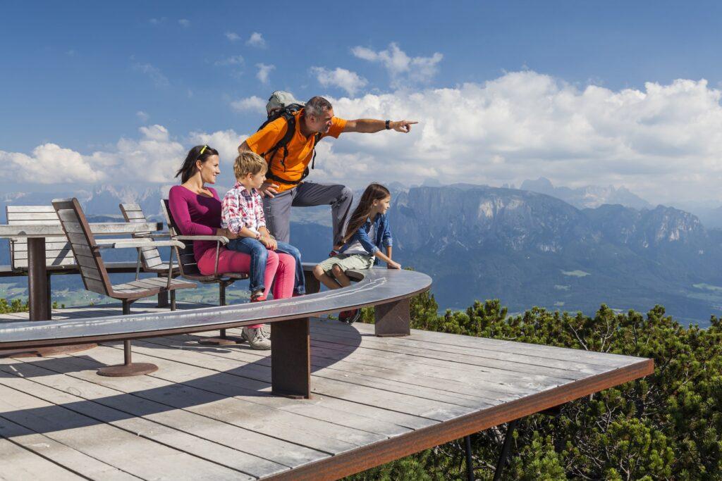 Eine vierköpfige Familie genießt von einer Holzterrasse aus den malerischen Blick auf die Berge Südtirols. Der Vater zeigt auf die Landschaft und schafft so einen Moment voller Staunen und Abenteuerlust.