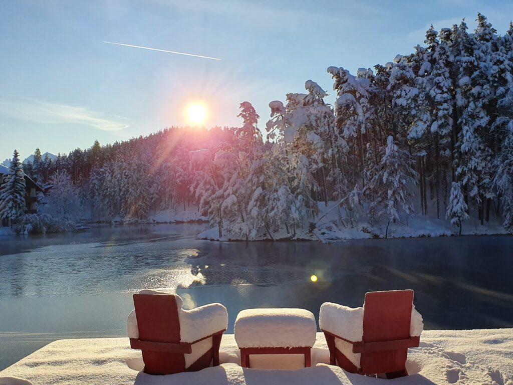 Schneebedeckte Stühle stehen bei Sonnenaufgang vor einem ruhigen, zugefrorenen See am Hotel Weihrerhof in Südtirol, der von schneebedeckten Bäumen umgeben ist. Die strahlende Sonne taucht die Winterlandschaft in ein warmes Licht.