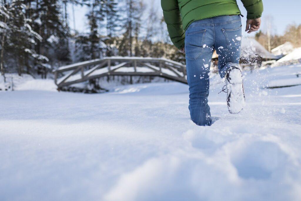 Eine Person in grüner Jacke und Jeans läuft durch tiefen Schnee auf eine Holzbrücke in einem verschneiten Wald zu und vermittelt dabei ein Gefühl von Abenteuer und winterlicher Ruhe in Südtirol.