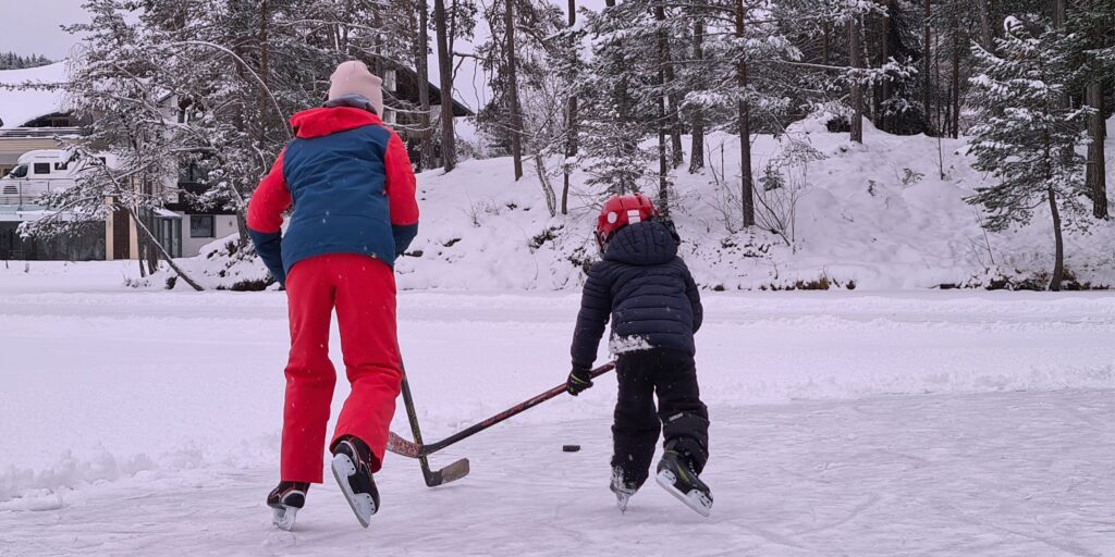 Ein Kind und ein Erwachsener in Winterkleidung spielen Eishockey auf dem zugefrorenen Wolfsgrubensee, umgeben von schneebedeckten Bäumen, und vermitteln so eine fröhliche Winterszene in Südtirol.