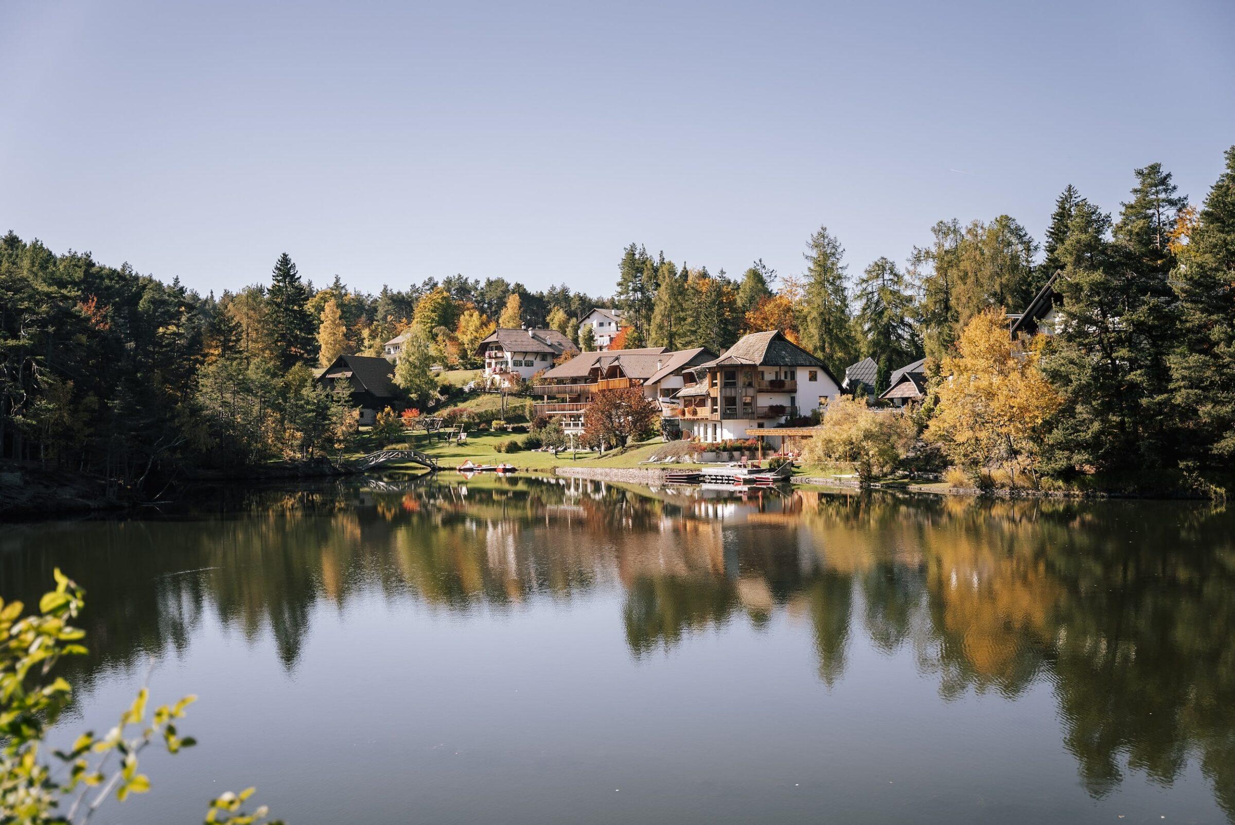 Das ruhige Hotel Weihrerhof am Seeufer in Südtirol mit seinen gemütlichen Gebäuden, umgeben von üppigen Herbstbäumen, die sich im ruhigen Wasser unter einem klaren blauen Himmel spiegeln, strahlt Ruhe und Frieden aus.
