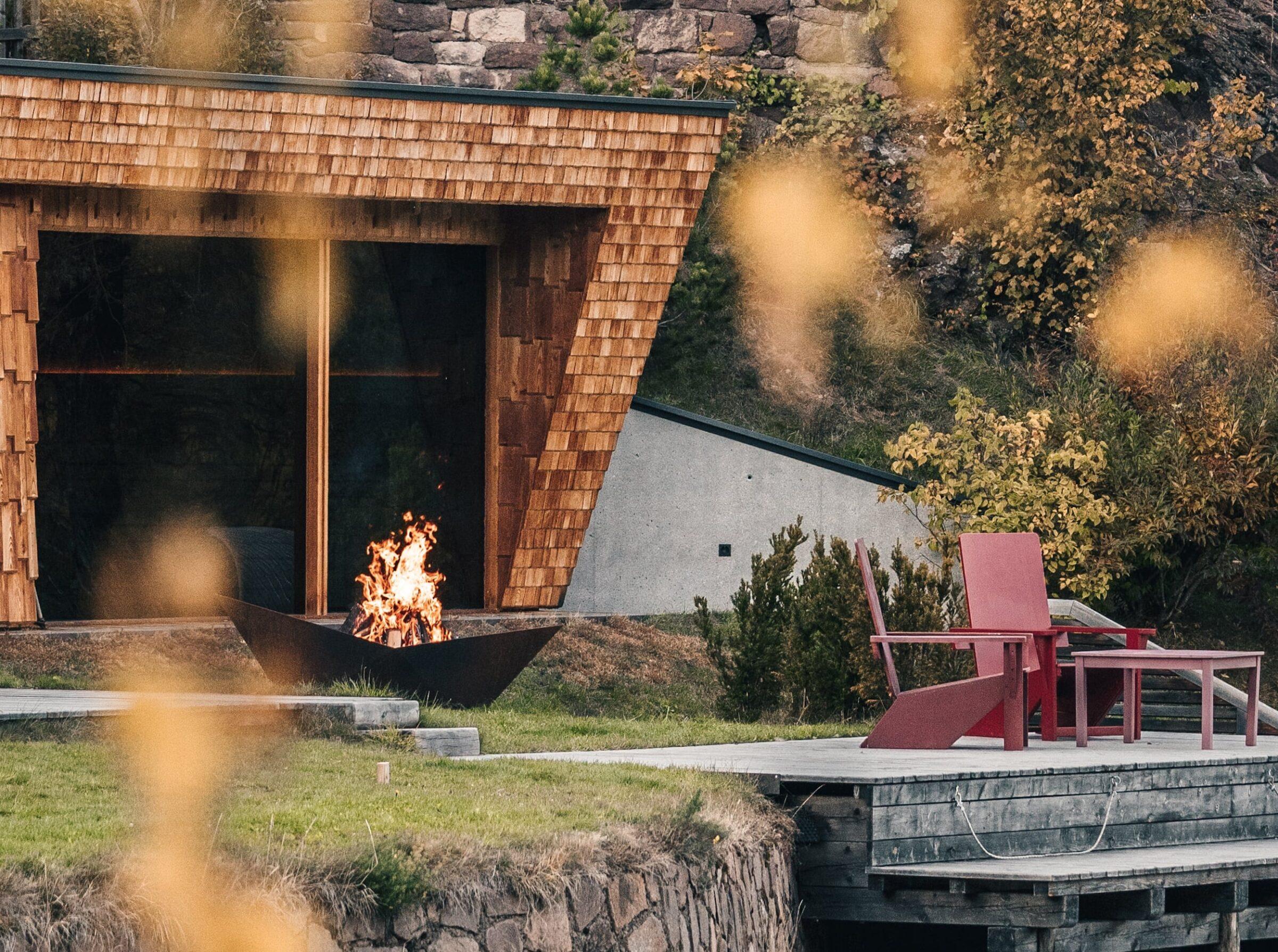 Moderne Bootshaussauna mit Holzfassade und Kamin auf einer grasbewachsenen Böschung im nachhaltigen Hotel Weihrerhof in Südtirol. Rote Stühle auf einem Steg, umgeben von Herbstlaub, sorgen für eine gemütliche und friedliche Atmosphäre.