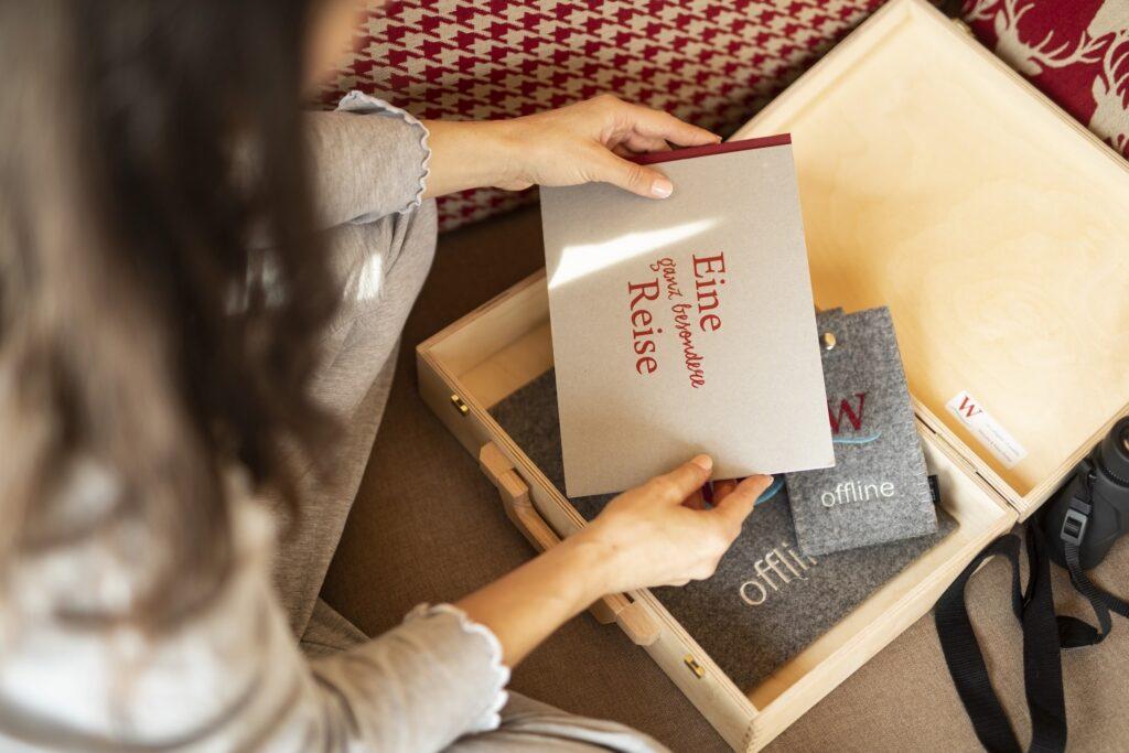 Eine Frau öffnet eine Holzkiste und hält ein Büchlein mit dem Titel „Eine ganz besondere Reise“ in der Hand. Die Szene wirkt gemütlich, mit sanfter Beleuchtung und rot-weiß gemusterten Kissen in der Nähe. Es ist das Digital Detox Angebot des nachhaltigen Hotels Weihrerhof in Südtirol