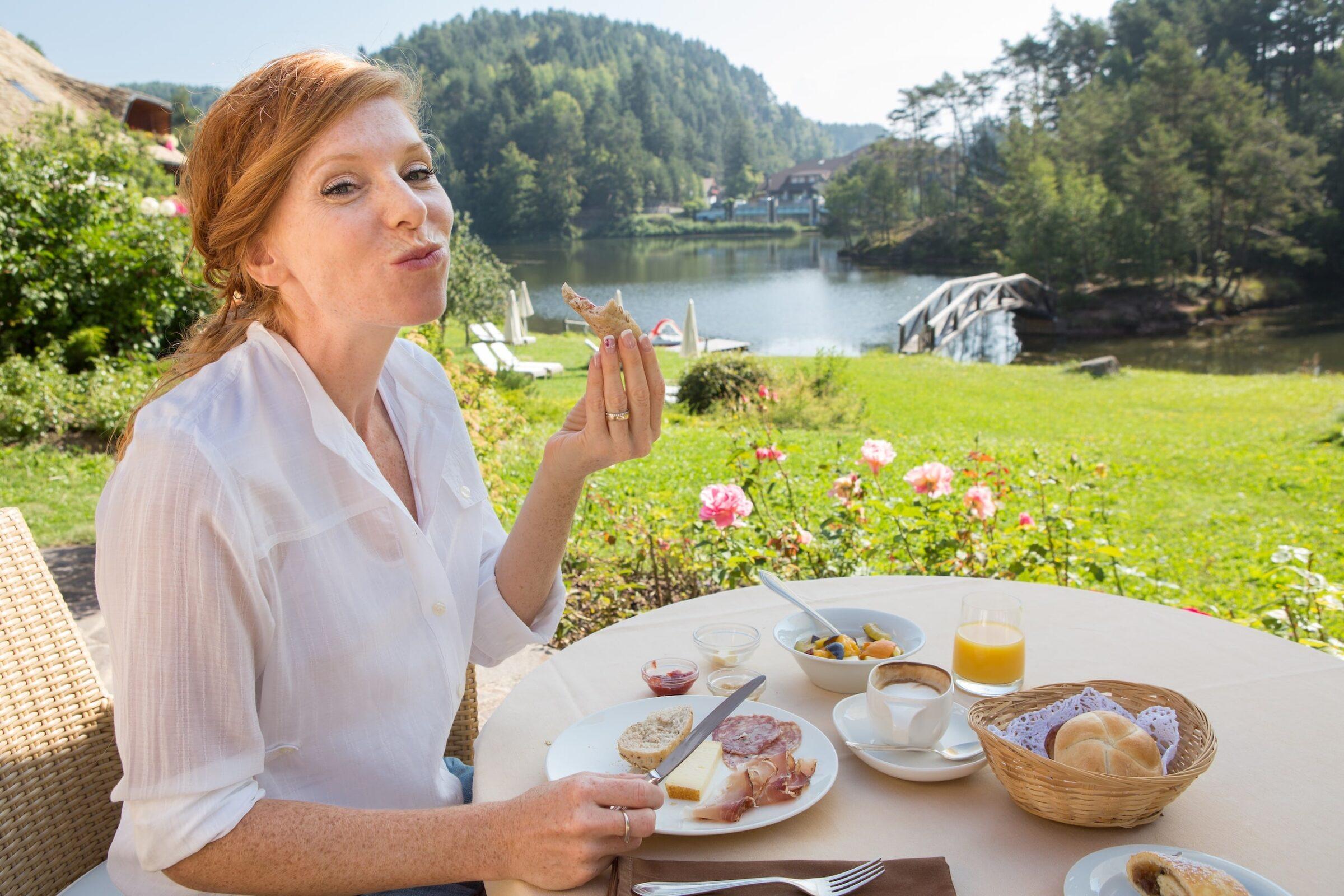 Frau genießt ihr Frühstück im Freien am ruhigen Wolfsgrubensee auf der Terrasse des nachhaltigen Hotels Weihrerhof, umgeben von Grün und rosa Blumen. Auf dem Tisch stehen Brötchen, Saft und Frühstückszutaten.
