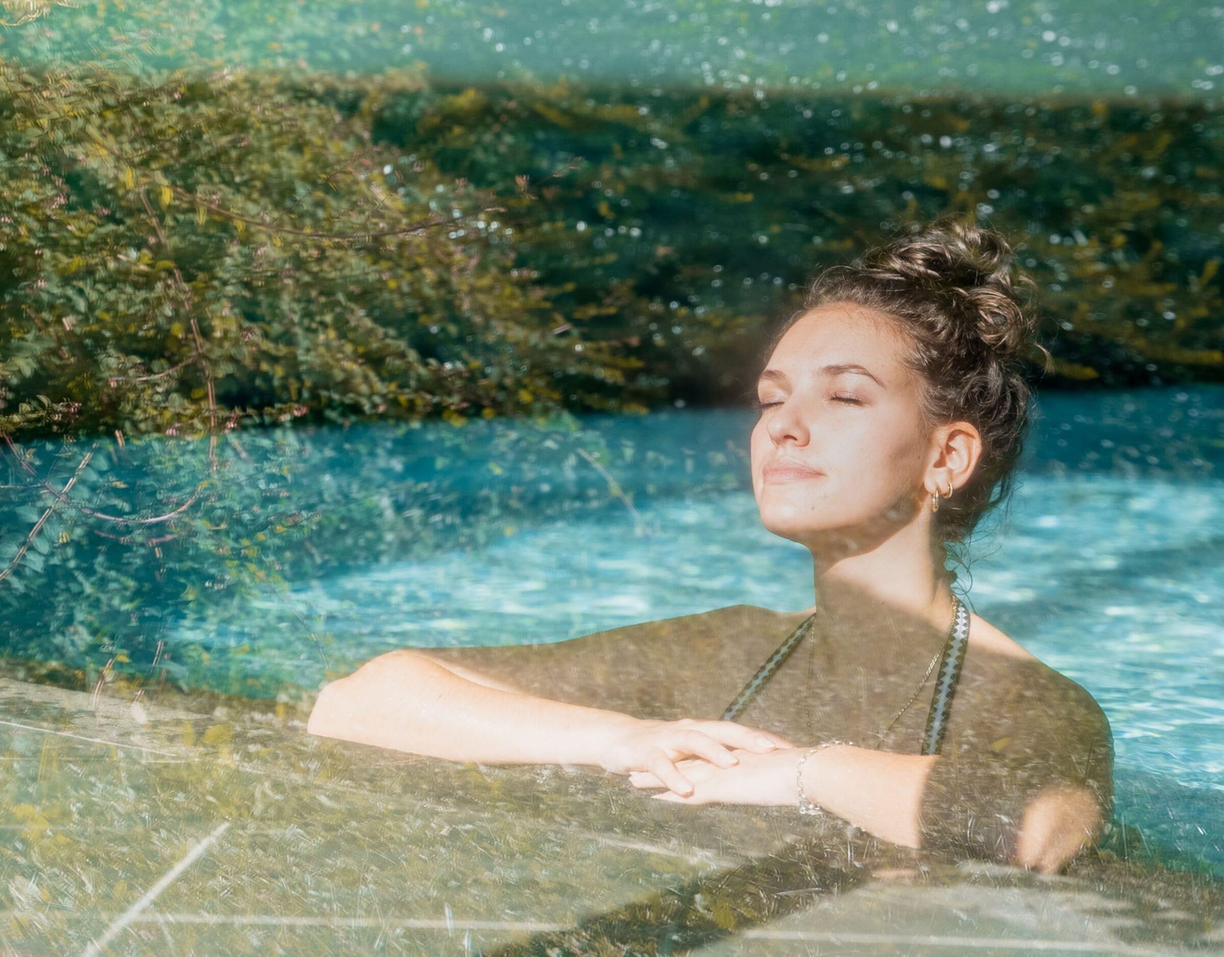 Eine Frau chillt mit geschlossenen Augen in einem sonnigen Pool im nachhaltigen Hotel Weihrerhof in Südtirol, ihr Gesicht strahlt Ruhe aus. Üppiges grünes Laub spiegelt sich im Wasser und schafft eine ruhige, verträumte Atmosphäre.