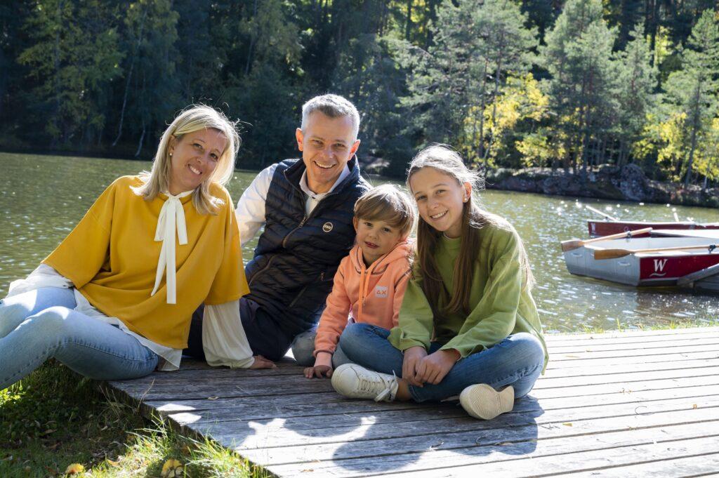 Die vierköpfige Inhaberfamilie Pichler des Hotels Weihrerhofs sitzt auf einem Holzsteg an einem sonnigen, ruhigen See. Sie lächeln, umgeben von üppig grünen Bäumen und zwei Ruderbooten im Hintergrund.