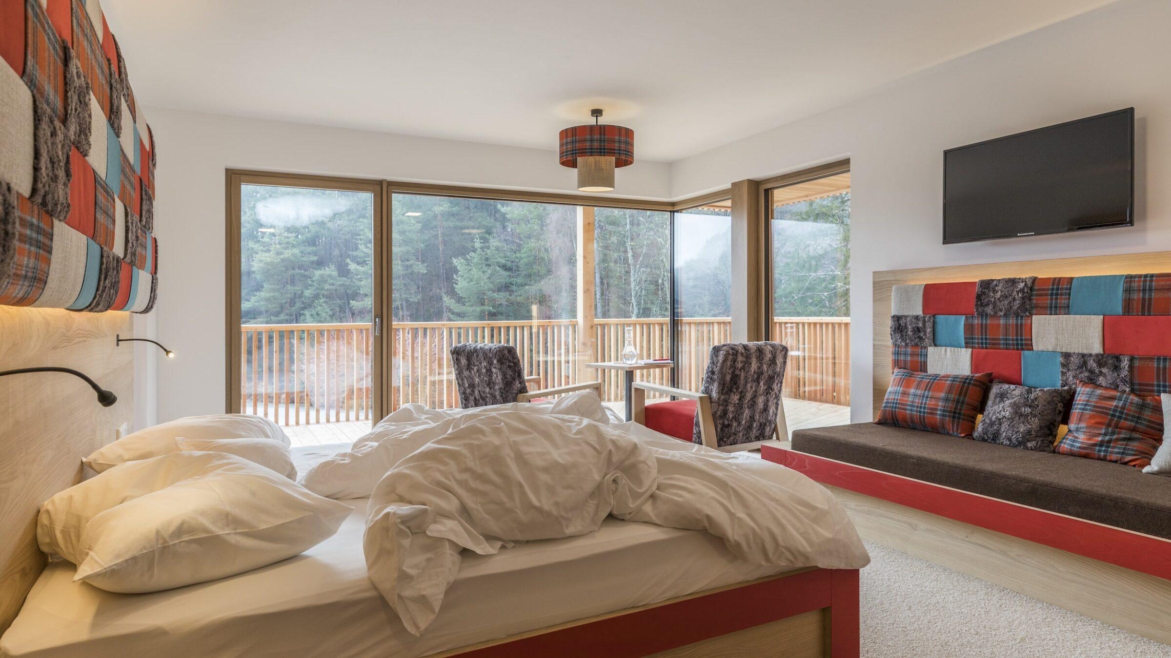 Modernes Hotelzimmer im nachhaltigen Hotel Weihrerhof in Südtirol mit einem ungemachten Bett, karierten Akzenten und großen Fenstern mit Blick auf den Wald. Gemütliche Sitzecke, beige Wände und eine ruhige Atmosphäre.