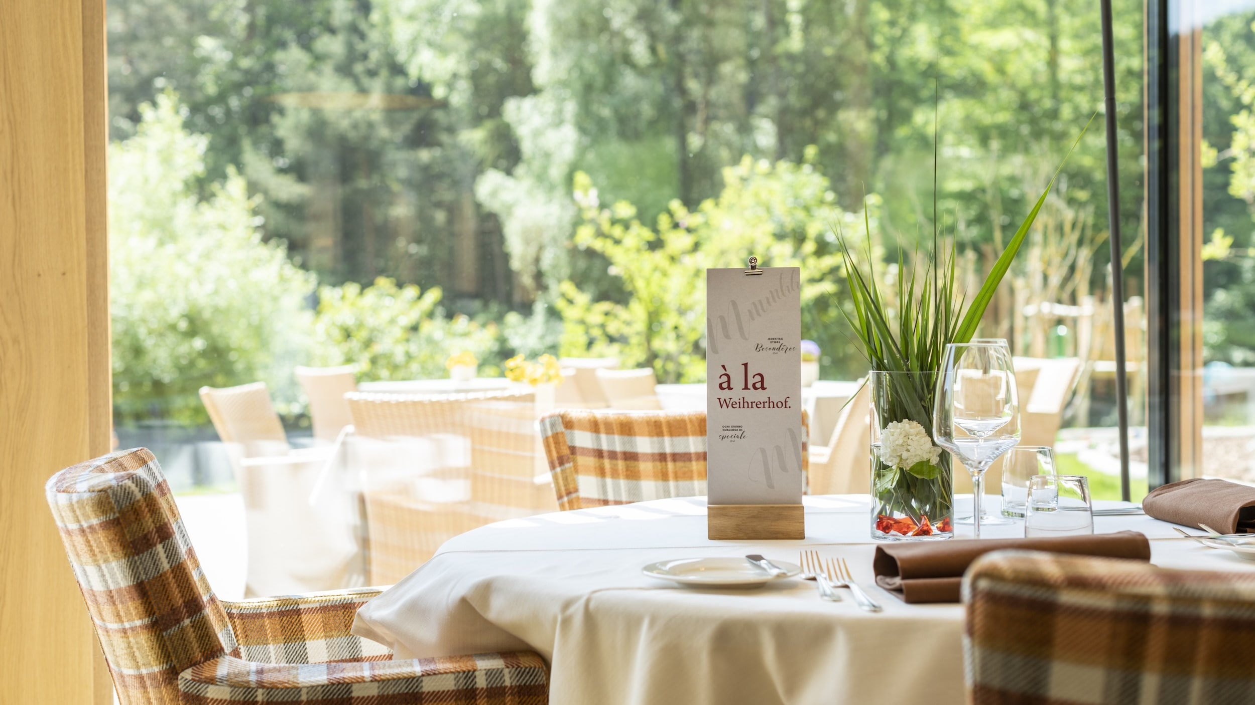 Ein schicker Restauranttisch für zwei Leute am großen Fenster mit Blick auf den grünen Garten im nachhaltigen Familienhotel Weihrerhof. Auf dem Tisch liegen die Speisekarte, eine Vase mit Blumen und edles Geschirr, was eine ruhige und einladende Atmosphäre schafft.