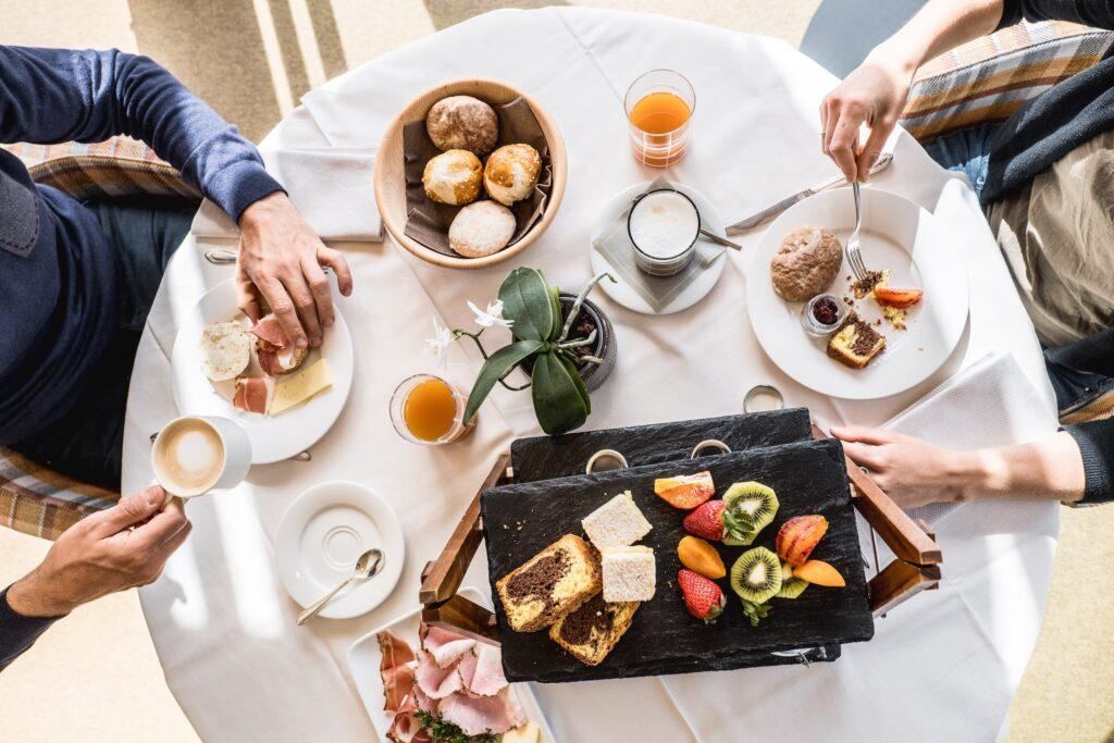 Luftaufnahme einer Frühstücksszene im nachhaltigen Hotel Weihrerhof in Südtirol an einem runden Tisch mit Gebäck, Obst, Kaffee und Saft. Zwei Personen sitzen dort und genießen gemeinsam eine gemütliche Mahlzeit.