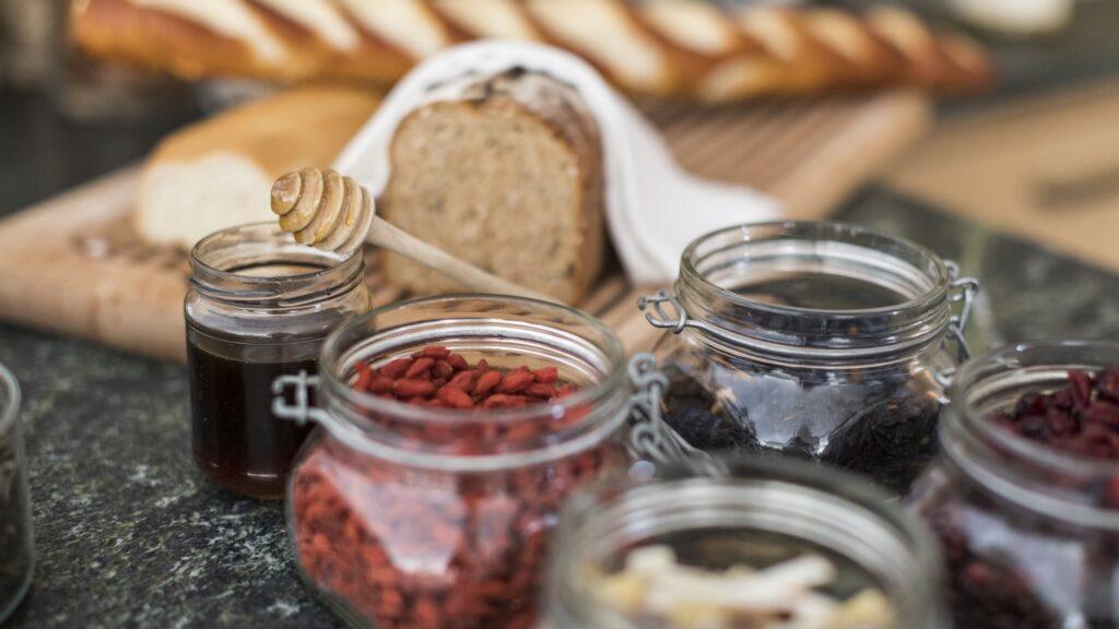 Eine rustikale Küchenszene mit Gläsern voller Trockenfrüchten und Honig im Fokus. Im Hintergrund frisches Brot und Baguettes auf einem Holzbrett. Warm und einladend.