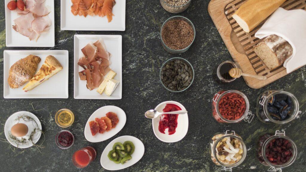 Luftaufnahme eines Frühstücksbuffets im familienfreundlichen und nachhaltigen Hotel Weihrerhof in Südtirol auf einem dunklen Tisch. Mit Tellern mit Aufschnitt, Brot, einem Ei, Obstscheiben, Samen und Gläsern mit Konfitüre. Gemütlich und einladend.