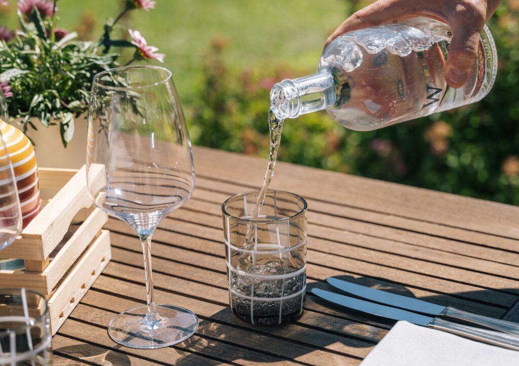 Eine Hand gießt Sprudelwasser aus einer durchsichtigen Flasche in ein Glas auf einem Holztisch. Ein Weinglas, Besteck und ein Korb mit Blumen sorgen für eine entspannte Atmosphäre im Freien.